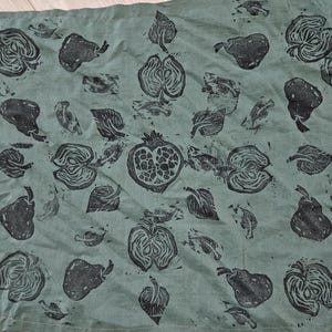 May include: A teal linen napkin featuring a repeating pattern of black fruit and leaf block prints. The design includes pears, apples, pomegranates, and various leaf shapes. The fabric has a slightly wrinkled texture.