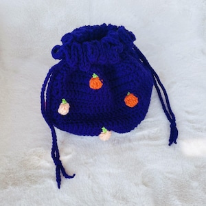 Puede incluir: Un bolso de ganchillo azul oscuro hecho a mano con cordón. El bolso está adornado con pequeñas aplicaciones de calabaza de color naranja y melocotón con tallos verdes. El borde superior está fruncido y el bolso tiene un cierre de cordón. El bolso está sobre una superficie blanca y esponjosa.