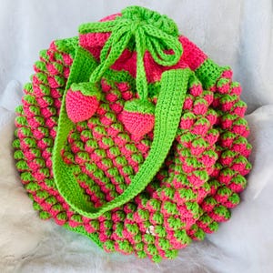 May include: A handmade crochet bag in a strawberry design. The bag is primarily pink with green accents, including the strap, drawstring, and strawberry details. The bag is round and textured, with a drawstring closure.