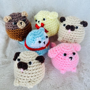May include: A collection of five small, round, crocheted plush toys. The group includes a brown bear, a pale yellow creature with a red bow, a light blue animal with a red collar, a beige pug with dark ears, and a pink creature with floppy ears.