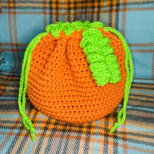 Op de afbeelding: Een gehaakte, pompoenvormige trekkoordtas van oranje en groen garen. De tas heeft een groen, gehaakt blad aan de bovenkant.
