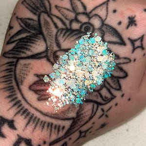 Peut inclure: Un tas de paillettes irisées dans les tons de bleu, vert et blanc. Les paillettes sont sur une main avec un tatouage noir détaillé d'un motif floral, d'un croissant de lune et d'étoiles.