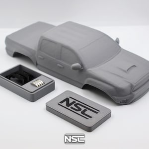 Può includere: Modello grigio stampato in 3D di un pick-up con cassone separato, componenti del motore e una piastra rettangolare con la scritta "NSC". Il modello è su una superficie bianca.