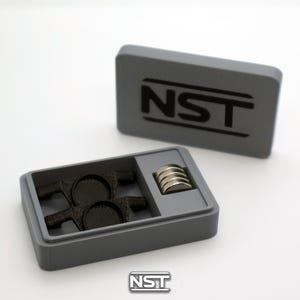 May include: A rectangular gray storage box with the text NST printed in black on the lid. The open box contains two black textured components with circular centers and a small stack of five metallic silver circular magnets in a side compartment.