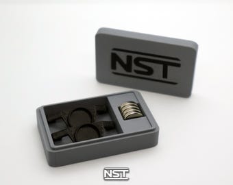 Op de afbeelding: Een rechthoekige grijze opbergdoos met de tekst NST in het zwart op het deksel. De open doos bevat twee zwarte, getextureerde onderdelen met cirkelvormige centra en een kleine stapel van vijf zilverkleurige, ronde magneten in een vakje.