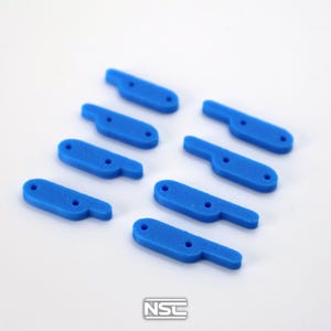 Pu&ograve; includere: Una collezione di otto componenti a forma di leva in plastica blu brillante. Ogni pezzo ha un'estensione a forma di manico e due piccoli fori. Gli oggetti sono disposti su una superficie bianca. Il logo "NSC" &egrave; visibile in basso a destra.