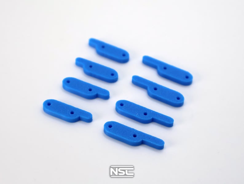 Pu&ograve; includere: Una collezione di otto componenti a forma di leva in plastica blu brillante. Ogni pezzo ha un'estensione a forma di manico e due piccoli fori. Gli oggetti sono disposti su una superficie bianca. Il logo "NSC" &egrave; visibile in basso a destra.