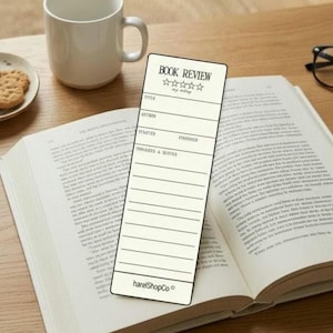 Può includere: Un segnalibro color crema per recensioni di libri con la scritta "BOOK REVIEW" e spazi per titolo, autore e note. È appoggiato su un libro aperto, con una tazza bianca e biscotti sullo sfondo.