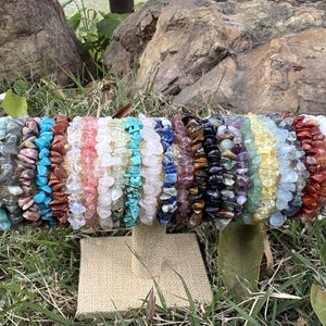 May include: A display of colorful gemstone chip bracelets. The bracelets are arranged on a beige stand, showcasing a variety of colors including blue, green, red, and purple. The background features grass and rocks.