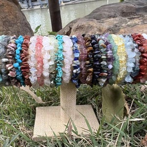 May include: A display of gemstone chip bracelets in various colors, including blue, green, pink, and brown. The bracelets are strung together and presented on a beige stand, set against a backdrop of greenery and water.