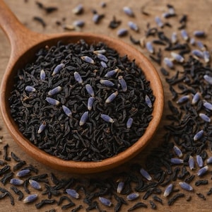 May include: A wooden spoon filled with black tea leaves and lavender buds. The tea is dark brown, and the lavender buds are a pale purple. The spoon sits on a wooden surface, with scattered tea and lavender around it.