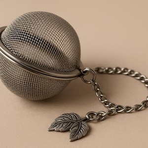 May include: A stainless steel tea infuser with a mesh ball and a chain. The chain ends with a decorative leaf charm. The infuser is designed for steeping loose leaf tea.