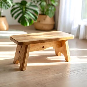 May include: A small wooden step stool, crafted from light-coloured wood. It features a rectangular top and four legs, showcasing a simple, minimalist design. The stool, approximately 30 cm in height, is suitable for use as a footrest or a small seat.