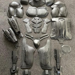 May include: A disassembled, metallic gray cosplay armor set, laid out on a gray surface. The set includes a helmet, chest and back pieces, shoulder pads, arm and leg guards, and other components. The armor has a futuristic, mechanical design.