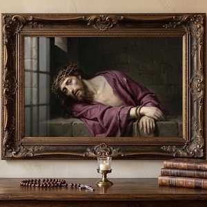 Pode incluir: Pintura emoldurada de um homem com coroa de espinhos e túnica roxa. A moldura ornamental marrom escuro envolve a obra de arte. Uma vela acesa, um rosário e livros empilhados estão sobre uma mesa de madeira sob a pintura.