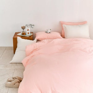 Pode incluir: Uma capa de edredom rosa com um travesseiro branco e um travesseiro rosa em uma cama. A cama é feita com um lençol branco e um cobertor rosa. Há uma pequena mesa de madeira com um vaso de flores e uma xícara de café em cima.