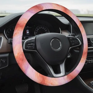 Op de afbeelding: Een stuurwielhoes met een aquarelontwerp in tinten oranje, roze en paars. De hoes is geïnstalleerd op een zwart stuurwiel. Het dashboard en het interieur van de auto zijn zichtbaar op de achtergrond.
