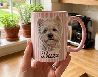 May include: A pink and white striped ceramic mug featuring a West Highland White Terrier portrait and the name "Buzz". The mug has a pink handle and rim. A hand is holding the mug.