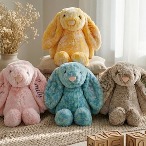 Pode incluir: Quatro coelhos de pelúcia em tons de amarelo, rosa, azul e castanho. Os coelhos têm orelhas longas e caídas e detalhes bordados. Um coelho tem o nome "Emily" bordado na orelha. Os coelhos estão sentados numa superfície estampada.
