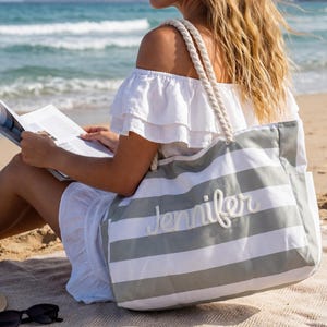 Può includere: Una grande borsa da spiaggia a righe orizzontali grigie e bianche. La borsa ha un manico in corda e il nome "Jennifer" ricamato sul davanti. La borsa è su una spiaggia sabbiosa.