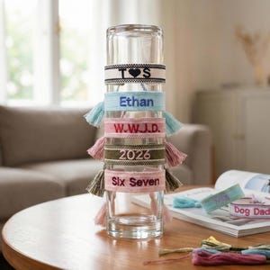 Könnte beinhalten: Ein klares Glas hält einen Stapel personalisierter, gestickter Armbänder in verschiedenen Farben. Die Armbänder zeigen Namen, Initialen und ein Jahr. Zusätzliche Armbänder liegen auf einer Zeitschrift, und Stickgarn liegt auf dem Tisch.