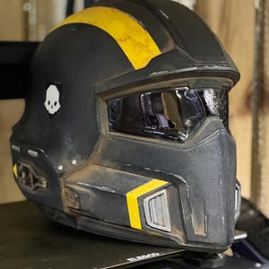 以下が含まれることがあります： 黄色いアクセントとスカルエンブレムが付いた黒いヘルメット。ヘルメットにはバイザーが付いており、頑丈で使い古された外観です。コスプレやコスチューム用と思われる。