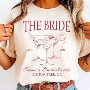 May include: A cream-coloured t-shirt with a burgundy graphic. The graphic features the text "THE BRIDE" above three martini glasses and the text "Emma's Bachelorette, Joshua Tree, CA". The year "2026" is also printed on the shirt.