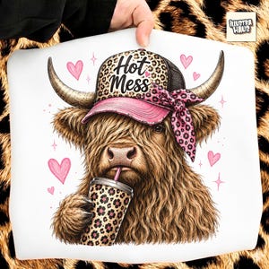 May include: A white t-shirt featuring a graphic of a brown Highland cow wearing a leopard print baseball cap with the text Hot Mess and a pink leopard print bow. The cow holds a leopard print cup with a pink straw, surrounded by pink hearts.