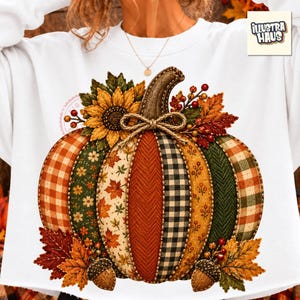 May include: A white sweatshirt featuring a large graphic of a pumpkin composed of various fabric patterns including orange gingham, green floral, autumn leaves, herringbone, and black plaid. It is decorated with a sunflower, berries, and acorns.