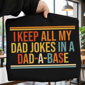 Könnte beinhalten: Schwarze Tragetasche mit dem Text "I KEEP ALL MY DAD JOKES IN A DAD-A-BASE" in orange, gelb und blaugrün. Der Text wird von orangefarbenen und blaugrünen horizontalen Linien umrahmt.