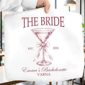May include: White tote bag with burgundy text reading "THE BRIDE". A burgundy martini glass with a bow is centered below the text. The text "EST. 2026" is on either side of the glass. The text "Emma's Bachelorette VARNA" is at the bottom.