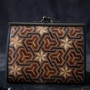 Pode incluir: Uma pequena carteira retangular com um padrão de mosaico de madeira geométrica em tons de castanho e bege. A carteira tem um fecho de metal escuro e uma borda de tecido preto. O design apresenta estrelas e hexágonos.