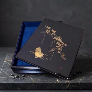 Puede incluir: Una caja rectangular negra con una tapa que presenta una ilustración dorada de un pájaro y una rama florida. El interior de la caja está forrado con una tela azul vibrante. La tapa está ligeramente abierta, revelando el forro azul interior.