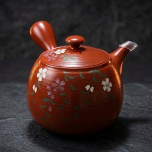 以下が含まれることがあります： 赤色のセラミックティーポット。取っ手、注ぎ口、蓋が付いています。ティーポットは白と紫の花柄と緑の葉で装飾されています。ティーポットは濃い灰色の表面に置かれています。