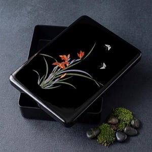 Può includere: Una scatola laccata nera con un coperchio con un motivo floreale con fiori arancioni e foglie verdi e due farfalle bianche. La scatola è aperta, rivelando l'interno. Una piccola composizione di muschio e pietre è accanto alla scatola.
