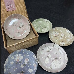 May include: Five ceramic plates with floral patterns in pink, green, beige, blue, and lavender. The plates are arranged on a dark surface, with one set inside a woven basket. The plates feature white and pink flower designs. The plates are decorated with white and pink flowers.