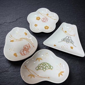 May include: Set of four white ceramic dishes in various shapes: a flower, a triangle, and two organic forms. Each dish features gold, red, and green decorative line art and floral accents. Ideal for serving small portions or as decorative pieces.