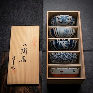 Puede incluir: Un juego de cinco cuencos de cerámica decorativos con intrincados diseños florales y paisajísticos en azul y blanco. Los cuencos se exhiben en una caja de madera con una tapa de madera a juego. La caja tiene caracteres japoneses.