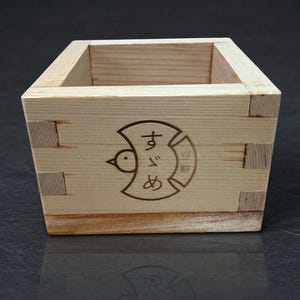 May include: A small, square wooden box with dovetail joints. The natural wood grain is visible. A circular emblem with a bird and Japanese text is etched on the front. The box is likely for serving or displaying small items.