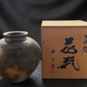 May include: A dark brown, round ceramic vase with a wide opening, next to a wooden box. The vase has a textured surface and a slightly lighter brown base. The wooden box has Japanese characters written on it.
