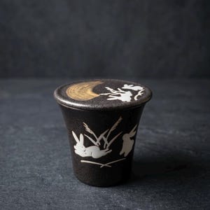 以下が含まれることがあります： 濃い茶色の陶器製カップと蓋のセット。白いウサギと背の高い草のイラストが描かれています。蓋には金色の三日月と白いウサギのモチーフがあしらわれています。表面全体に斑点のある質感の仕上げが施されています。