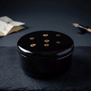 以下が含まれることがあります： 黒い漆塗りの丸い容器。蓋には7つの金色のリング状の装飾が施されています。容器は暗く、質感のある表面に置かれています。ミニマルでエレガントなデザインで、洗練された美しさを醸し出しています。