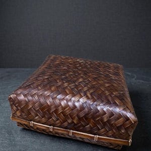 Puede incluir: Una caja rectangular con tapa hecha de tiras de bambú trenzado de color marrón oscuro. La tapa presenta un patrón de tejido en espiga diagonal apretado, mientras que la base está reforzada con un marco de bambú horizontal liso.