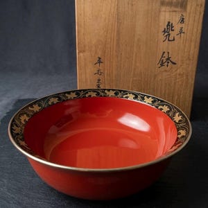 May include: A red lacquered bowl with a black and gold decorative rim. The bowl is set in front of a wooden box with Japanese calligraphy. The bowl has a glossy finish and a simple, elegant design.