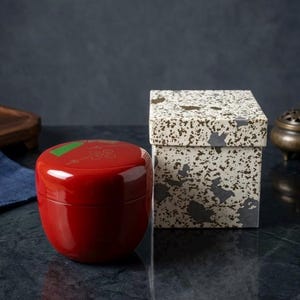 May include: A red, lacquered container with a green accent and floral design, next to a speckled box. The container has a rounded shape and a glossy finish. The box has a neutral color palette with dark gray and brown spots.