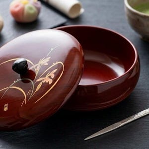 以下が含まれることがあります： 金色の花の装飾が施された、赤褐色の漆塗りの器。蓋は開いており、器の内側が見える。日本の菓子、抹茶碗、小さな銀色の道具が写っている。伝統的な日本の食文化を象徴するシーン。