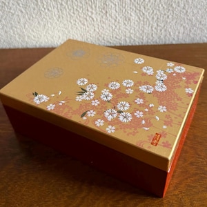 以下が含まれることがあります： 赤いベースと金色の蓋が付いた長方形の箱。蓋は白い桜の花とピンクのアクセントで装飾されています。この箱は日本の美学を持っています。