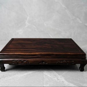 May include: A rectangular dark brown wooden display stand with a polished surface and visible wood grain. The stand features four curved legs with carved detailing along the front apron, providing a stable base for elevated presentation.