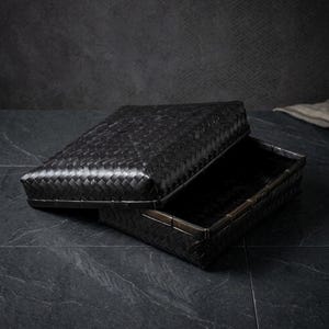 May include: A rectangular storage box crafted from woven black bamboo strips. The box features a matching removable lid that is partially lifted, revealing the hollow interior. The exterior surface displays a consistent diagonal weave pattern.