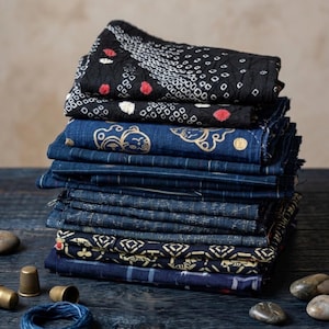 May include: A stack of folded fabric pieces in shades of black and indigo blue. The top black fabric features white geometric patterns and red accents. The blue fabrics display gold printed motifs, including cat figures and diamond shapes, alongside textured weaves.